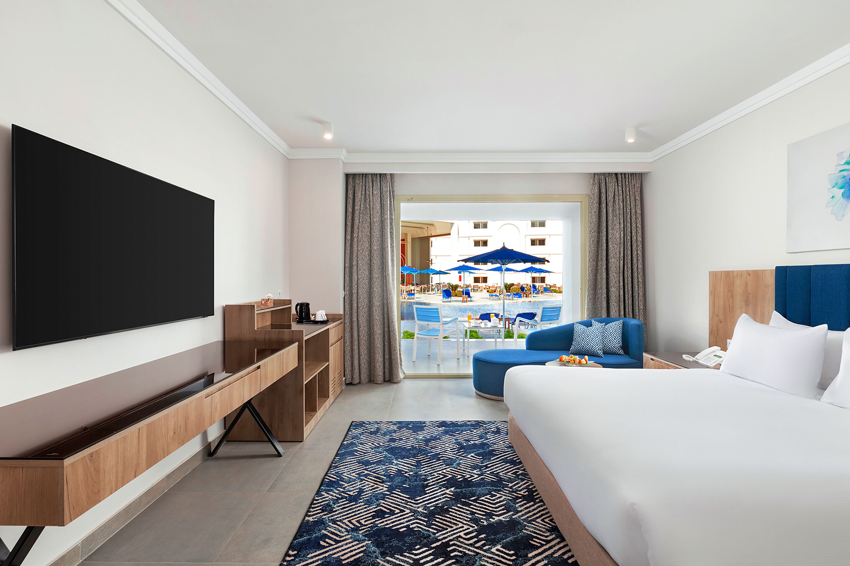 Una habitación de hotel moderna con una cama grande, un televisor de pantalla plana, una mesa consola de madera, un tapete azul estampado y vistas al área de la piscina.
