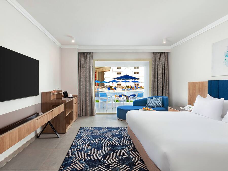 Una habitación de hotel moderna con una cama grande, un televisor de pantalla plana, una mesa consola de madera, un tapete azul estampado y vistas al área de la piscina.