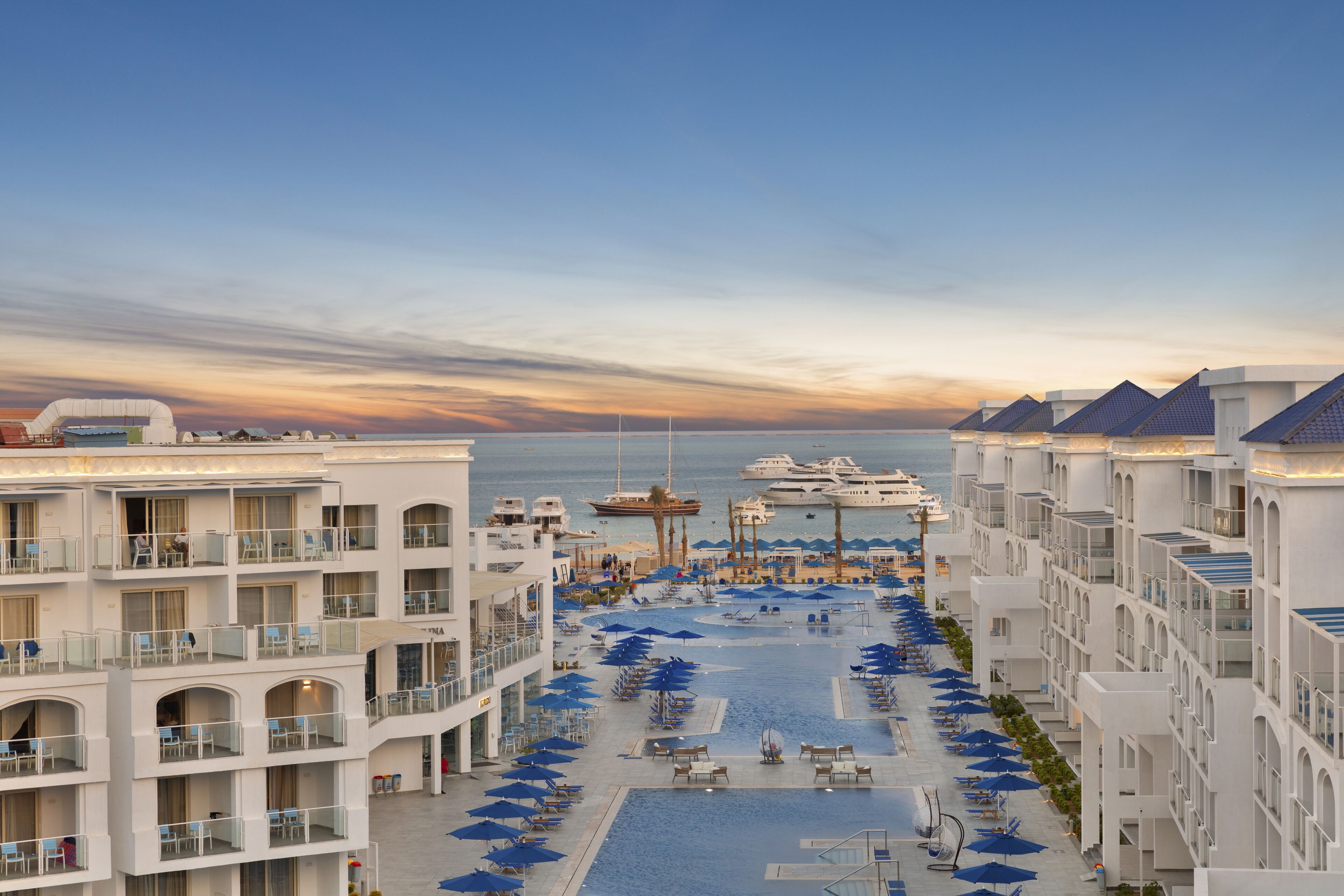 Ein Resort mit mehreren Balkonen, einem Poolbereich mit Sonnenschirmen und Blick auf das Meer.