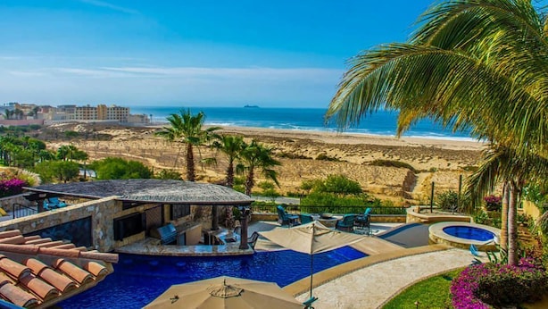 Zona de piscina con vista a la playa y al océano, rodeada de palmeras y sillones de descanso.