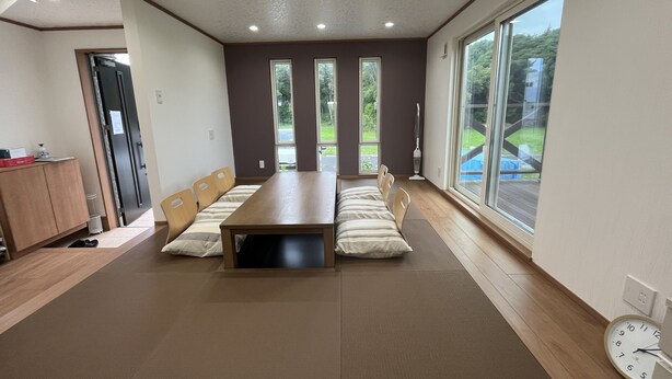 A spacious room with a long wooden table, cushioned seating, large windows, and a clock on the wall.