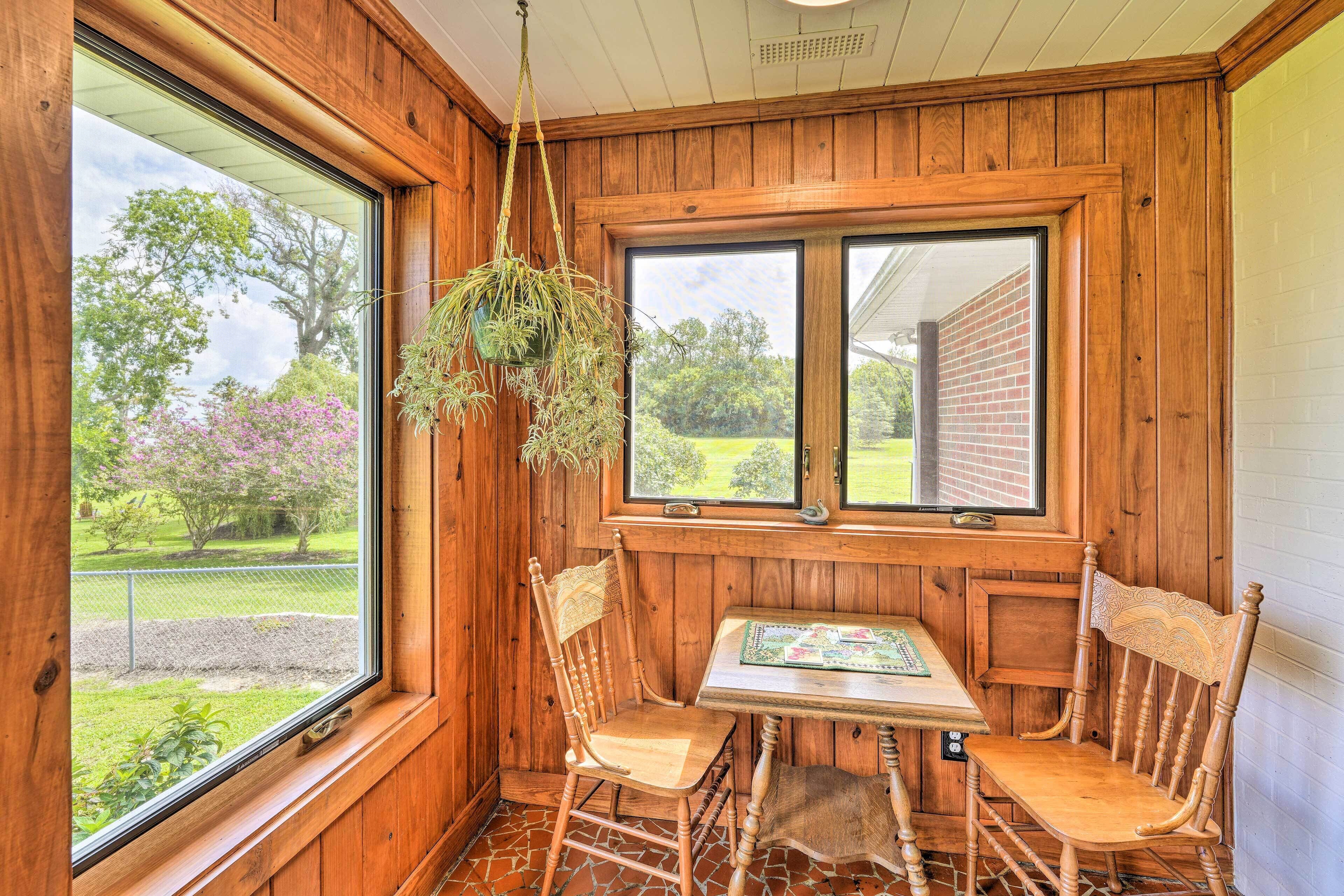 A wooden-paneled room with a table and two chairs, a hanging plant, and a view of a brick building and greenery outside.