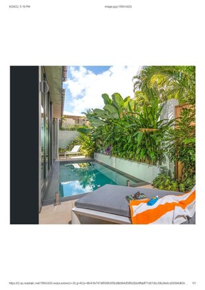 A modern outdoor pool area with a glass wall, lounge chairs, and lush tropical plants.