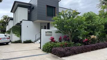 Une maison moderne à deux étages, avec une façade grise, un escalier apparent et un jardin bien entretenu où poussent différentes espèces de plantes.