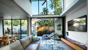 A modern living room with a sofa, coffee table, and TV. There is a large window with a view of greenery outside.