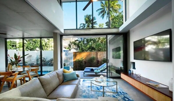 A modern living room with a sofa, coffee table, and TV. There is a large window with a view of greenery outside.