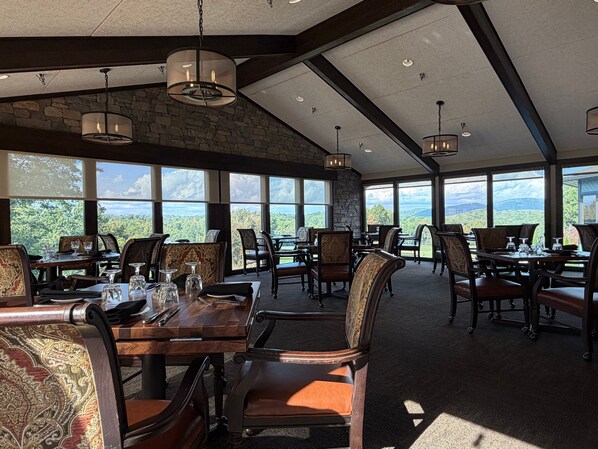 A dining area with tables and chairs, large windows, and a stone wall.