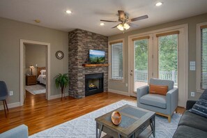 A living room with a fireplace, a ceiling fan, and large windows.