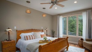 Un dormitorio con cama de madera, lámparas de noche, ventilador de techo y un ventanal con cortinas.