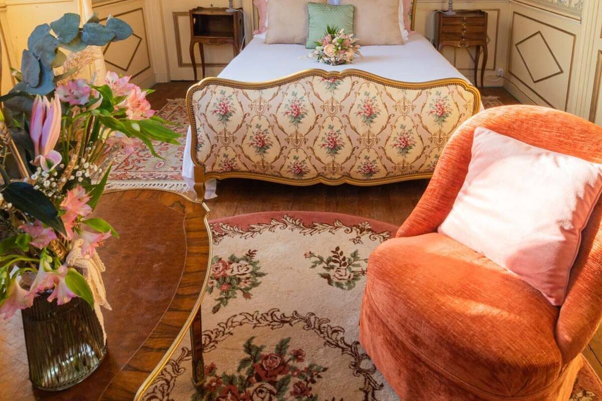 Quarto com papel de parede floral, cama com colcha florida, mesa de cabeceira de madeira, uma cadeira e um vaso com flores.