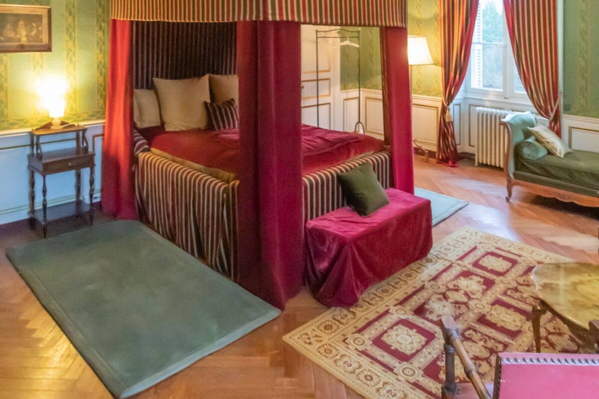 Uma cama com dossel, roupa de cama listrada em vermelho e branco, um banquinho estofado em verde e um tapete estampado sobre piso de madeira.