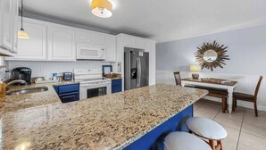 Una cocina con gabinetes blancos, encimera de granito, refrigerador y un comedor con mesa y sillas.