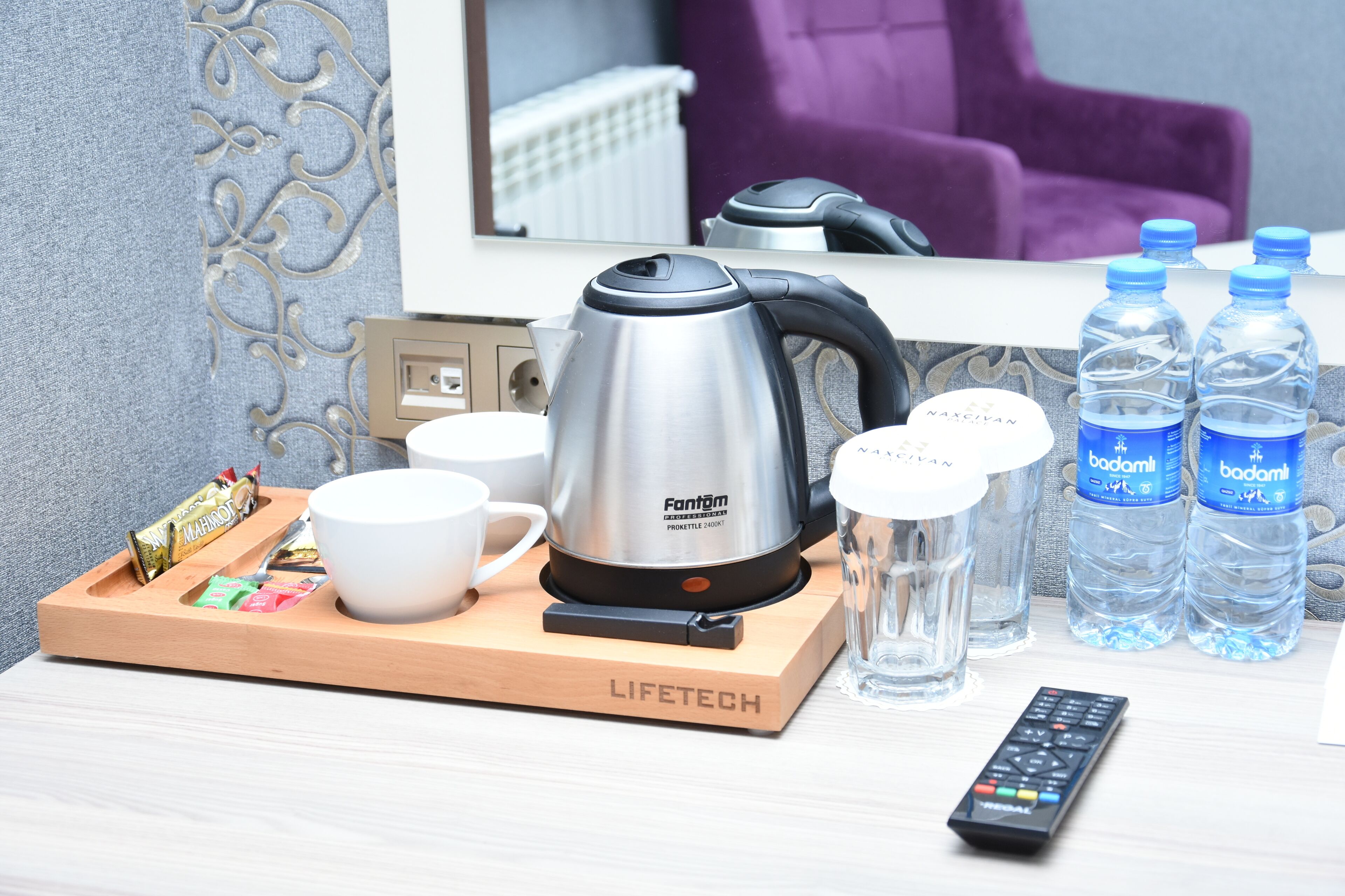 A hotel room with a kettle, cups, water bottles, a tray with snacks, and a remote control.