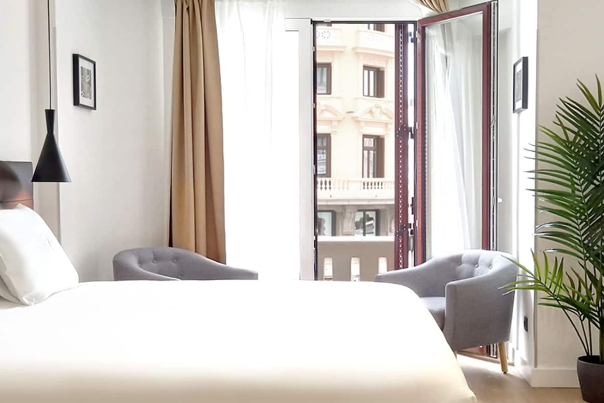 A hotel room with a large bed, two armchairs, a potted plant, and a view of a building through an open door.