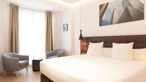 Habitación de hotel con una cama grande, dos sillones, una mesita auxiliar y un cuadro de paisaje urbano en la pared.
