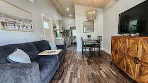A modern living room with a grey sofa, wooden flooring, a dining area with a table and chairs, and a TV on a wooden cabinet. - Surf Shack Tiny Home on Pinellas Trail w/ Heated Pool! Walk or Bike to Downtown Dunedin! (Dunedin)