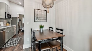 A modern kitchen with stainless steel appliances, a dining area with a wooden table and chairs, and a decorative chandelier. - Surf Shack Tiny Home on Pinellas Trail w/ Heated Pool! Walk or Bike to Downtown Dunedin! (Dunedin)