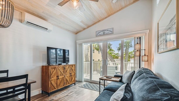 A modern living room with a wooden ceiling, a dark grey sofa, a wooden cabinet with a TV, and a sliding glass door leading to an outdoor area. - Surf Shack Tiny Home on Pinellas Trail w/ Heated Pool! Walk or Bike to Downtown Dunedin! (Dunedin)