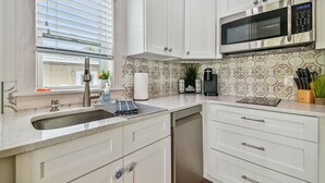 Una cocina moderna con armarios blancos, un microondas de acero inoxidable, un lavamanos con grifo y una ventana con persianas.