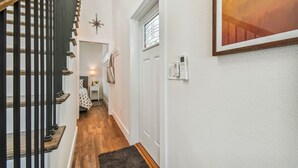 Un pasillo con una escalera de madera, una puerta blanca con ventana, un termostato en la pared y un cuadro enmarcado.