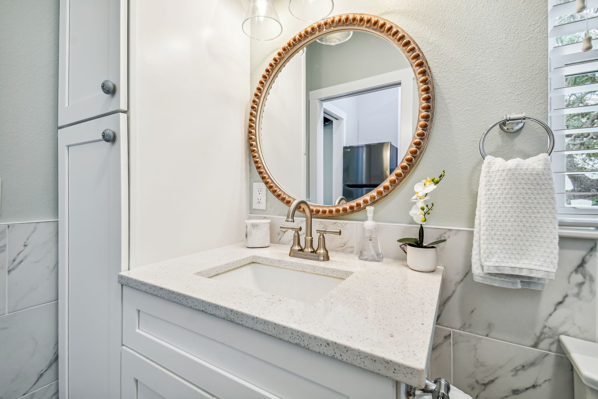 Un baño con un tocador blanco, un espejo redondo con un marco decorativo, un lavamanos y un perchero para toallas.
