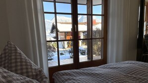 A hotel room with a large window offering a view of a snowy mountain and a building with a red roof. The room has two beds with checkered bedding.