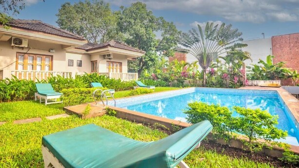 A swimming pool with lounge chairs, surrounded by greenery and a building with a tiled roof.