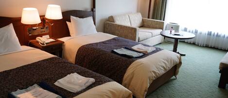 A hotel room with two beds, a chair, a small table, and a lamp.