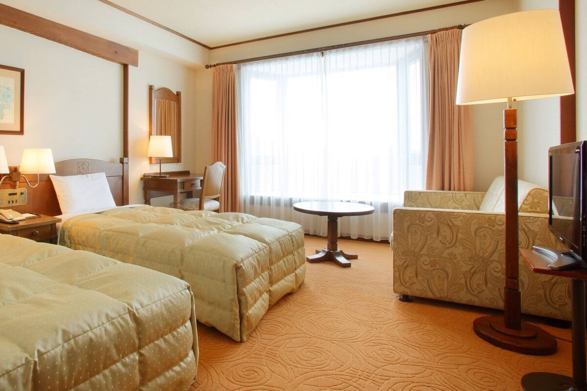 A hotel room with two beds, a desk, a chair, a TV, and a window with curtains.