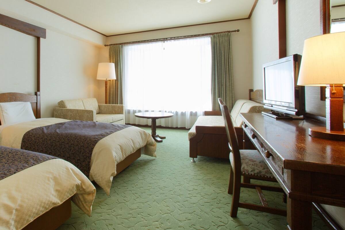 A hotel room with two beds, a desk with a television, a chair, and a lamp.