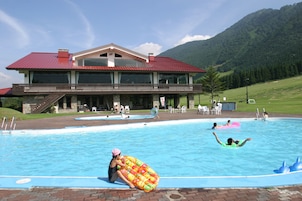 Una piscina con un edificio grande al fondo, una zona verde y montañas.