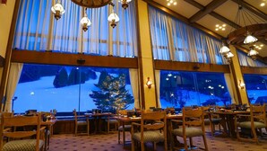 A dining area with wooden tables and chairs, large windows with curtains, and hanging light fixtures. - Hotel Onikoube (Osaki)