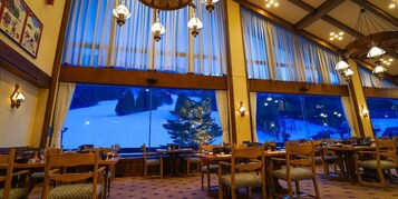 Un comedor con mesas y sillas de madera, ventanas amplias con cortinas, y lámparas colgantes.