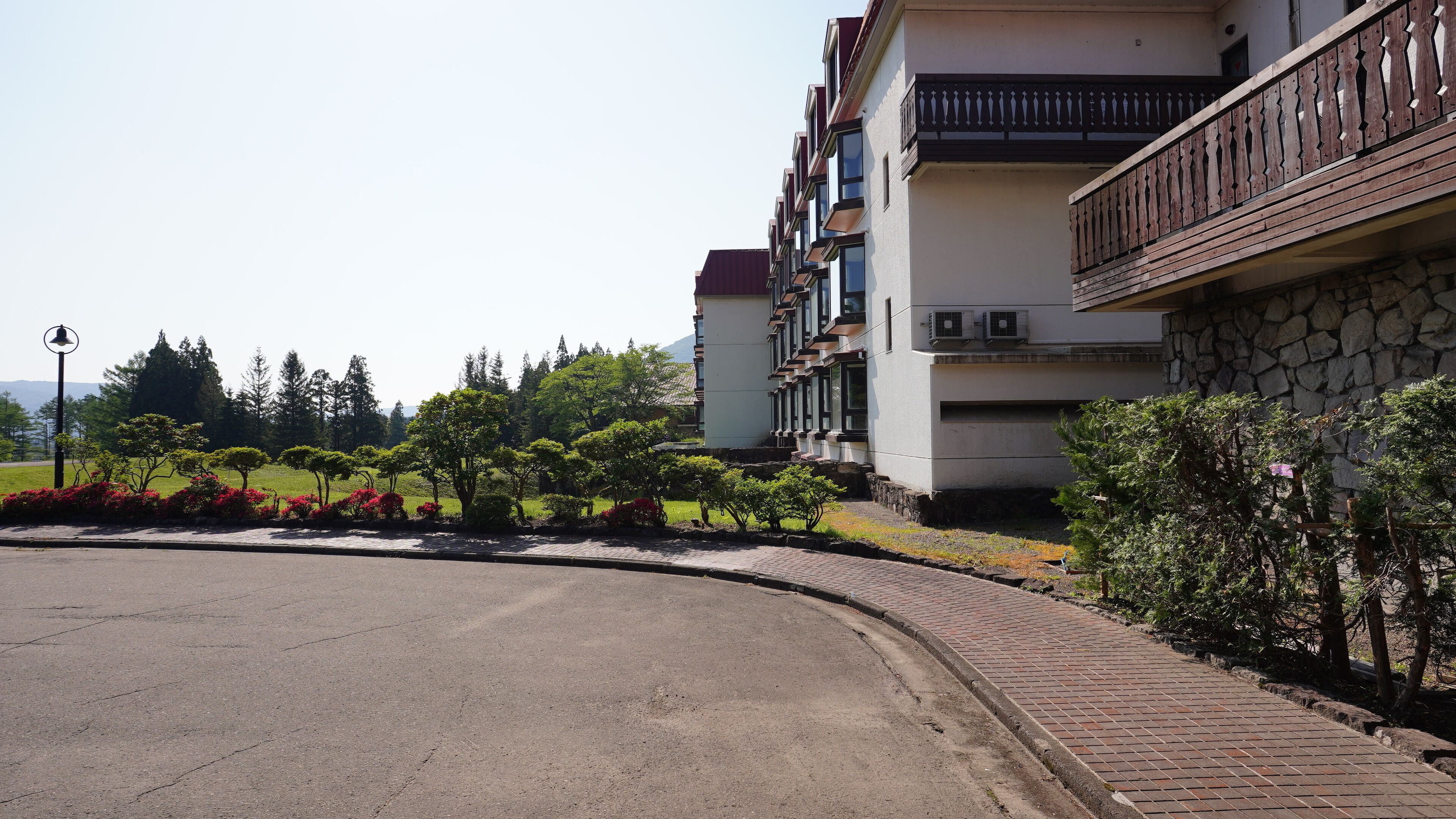 A multi-story building with balconies, a paved road, and landscaped areas with trees and shrubs.