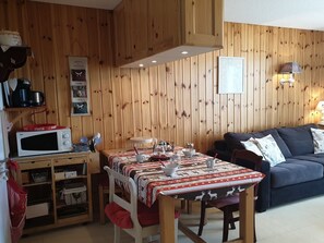 Tunnelmallinen huone, jonka seinät ovat puuta. Huoneessa on ruokapöytä, jonka päällä on pöytäliina, mikroaaltouuni ja tyynyillä varustettu sohva.