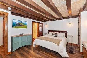 Un dormitorio con techo de madera, una cama con manta blanca y color crema, un televisor en la pared y un mueble pequeño.