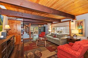 A spacious living room with a wooden ceiling, a staircase, a fireplace, and a large window.