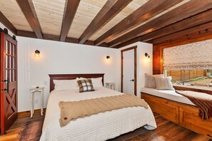 A bedroom with a wooden ceiling, a bed with white bedding, a bench with cushions, and a window with a view.