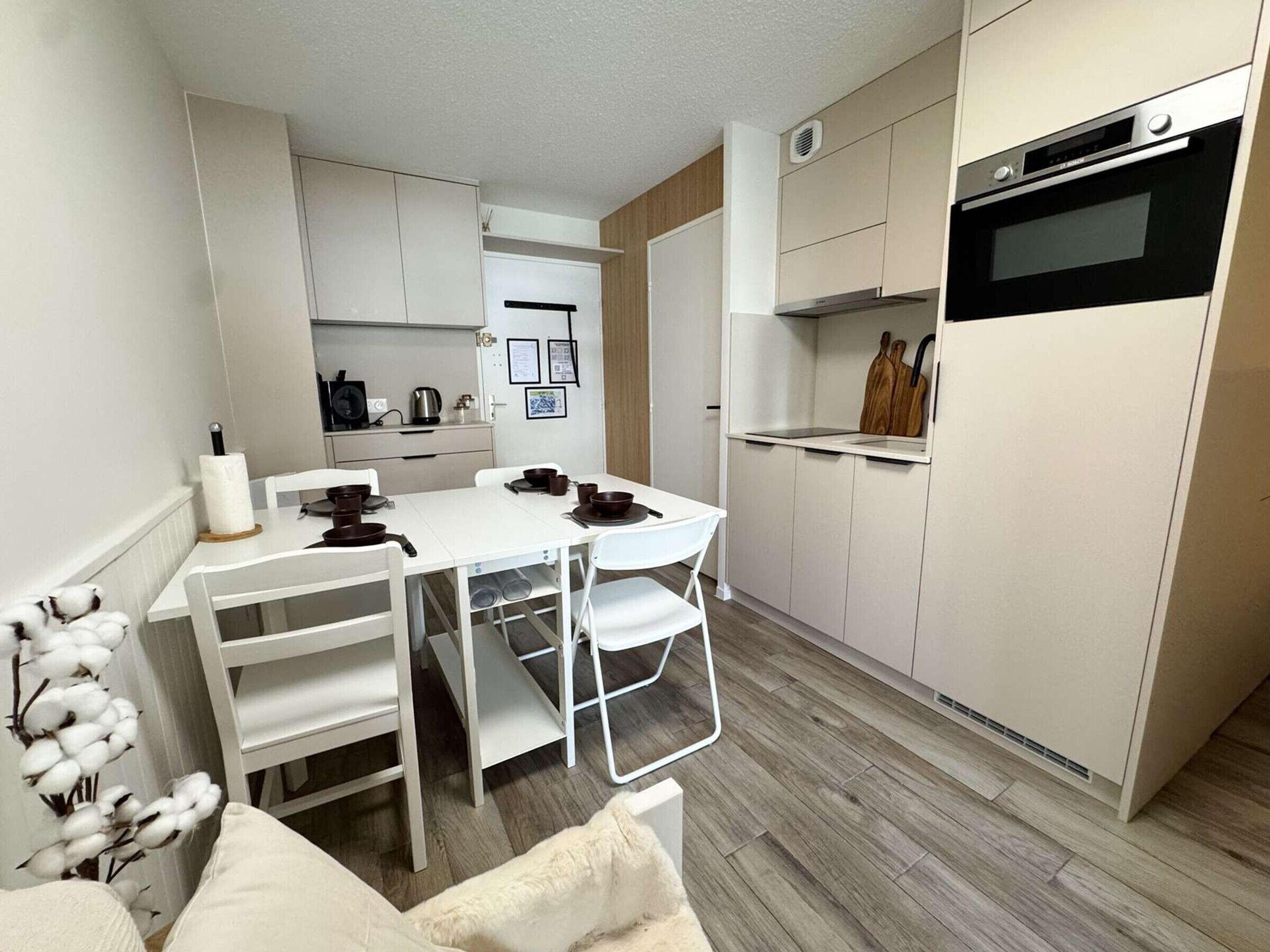 Una cocina compacta con muebles blancos, horno integrado y comedor con mesa y sillas blancas.