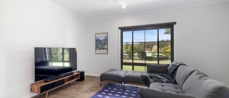 Una sala de estar moderna con un televisor de pantalla plana, un ventilador de techo, un ventanal con vistas a zonas verdes y una alfombra azul estampada.