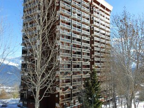 Um edifício de vários andares com inúmeras varandas, rodeado por árvores sem folhas e uma paisagem coberta de neve.