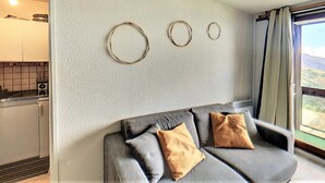 Un salon moderne avec un canapé gris et deux coussins, une cuisine en arrière-plan et une vue par la fenêtre.