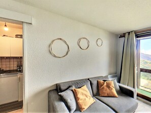 Sala moderna com sofá cinza e duas almofadas, cozinha ao fundo e vista pela janela.