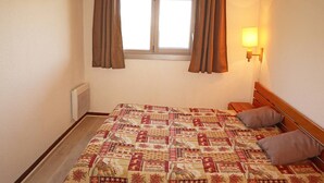 A bedroom with a bed, two pillows, a nightstand, a wall-mounted lamp, and a window with curtains.