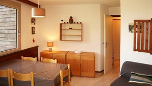 A cozy room with a wooden dining table, chairs, a sofa, a small cabinet with decorative items, and a window with blinds.