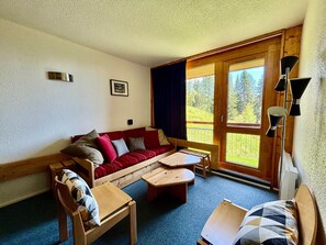 Um quarto aconchegante com um sofá de madeira, uma mesinha, uma cadeira e uma janela grande com vista para árvores.