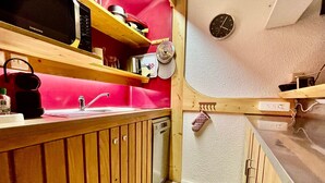 A compact kitchen with wooden cabinets, a microwave, a sink, and a clock on the wall.