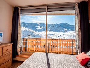 A room with a large window offering a mountain view, a wooden bench, a bed with a red pillow, and a wooden dresser. - Val Thorens - Apartment for 6, 3 Gold Flakes, 2 baths, near ski lifts (Val Thorens)