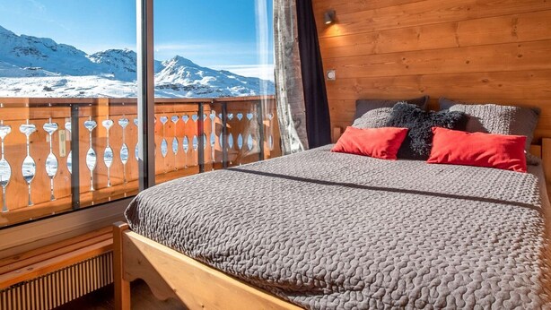 Un dormitorio con un ventanal grande, paredes de madera, una cama con ropa de cama gris y dos almohadas rojas.