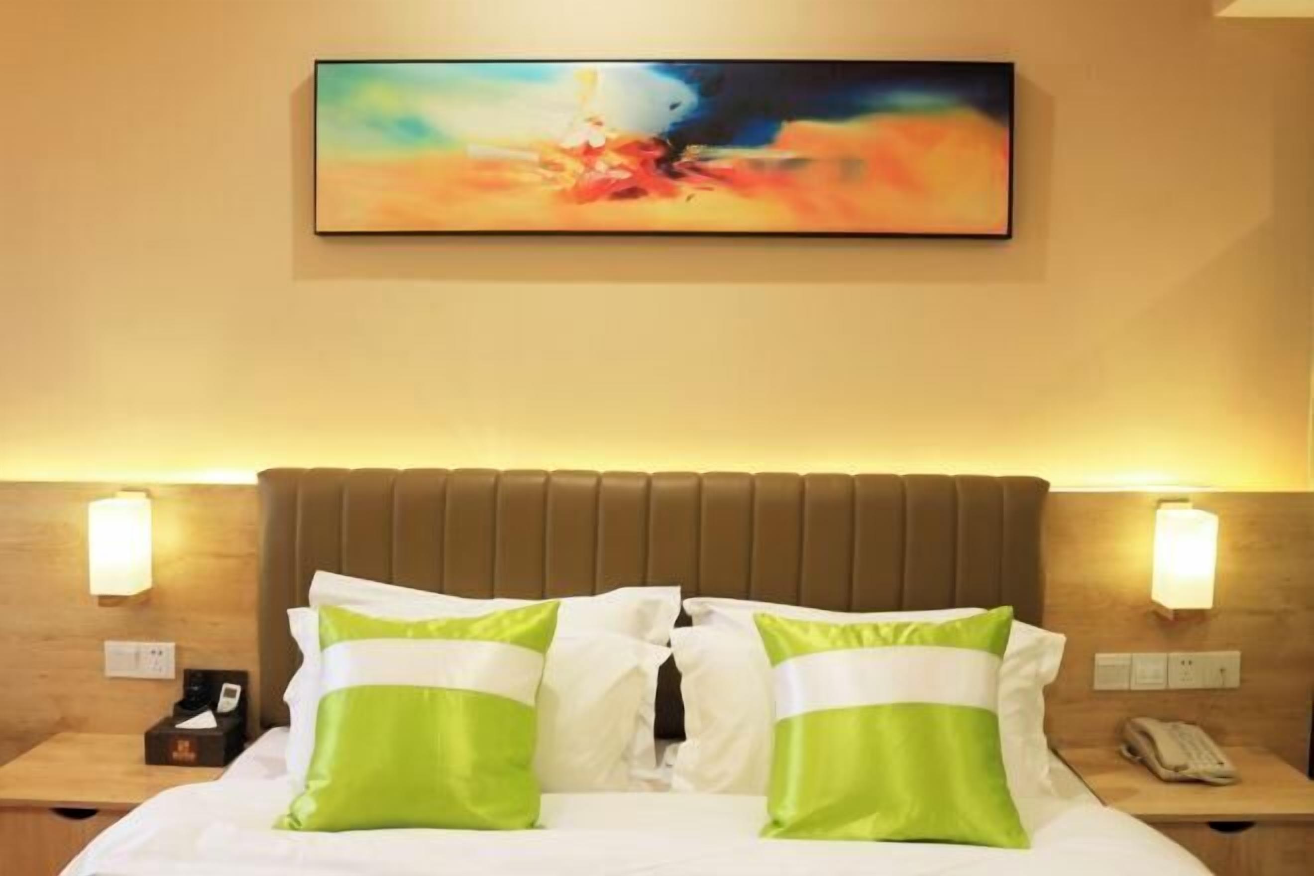 A hotel room with a bed, green and white pillows, a painting on the wall, and bedside lamps.
